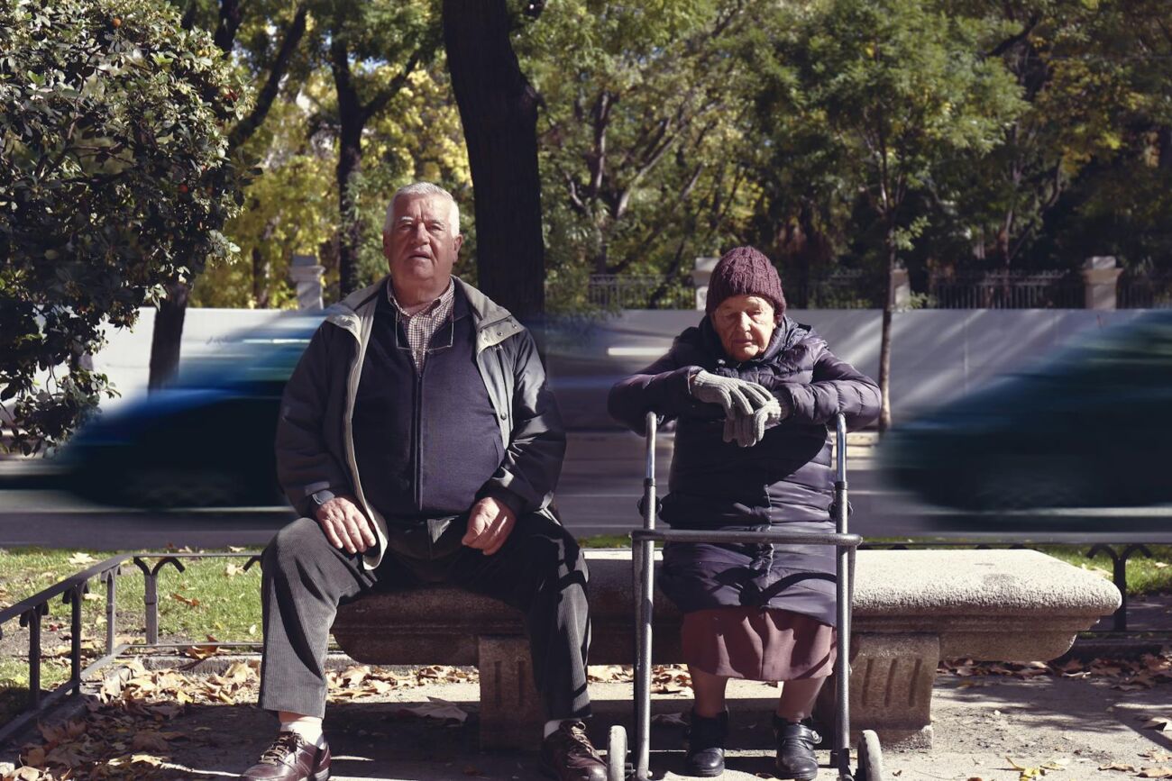 Senior couple enjoying a sunny day on a bench in Madrid park with blurred cars passing by.